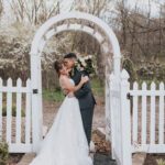 A bride and groom kiss in front of a white picket fence.