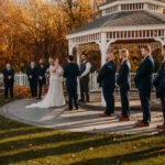 A bride and groom stand in front of a gazebo in the fall.