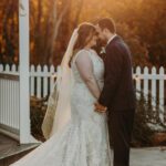 A bride and groom kissing in front of a white picket fence.