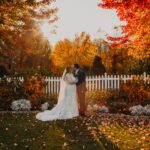 A bride and groom kissing in front of a white picket fence in the fall.