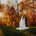 A bride and groom standing in a field with autumn leaves in the background.