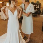 A bride is helping her mother put on her wedding dress.